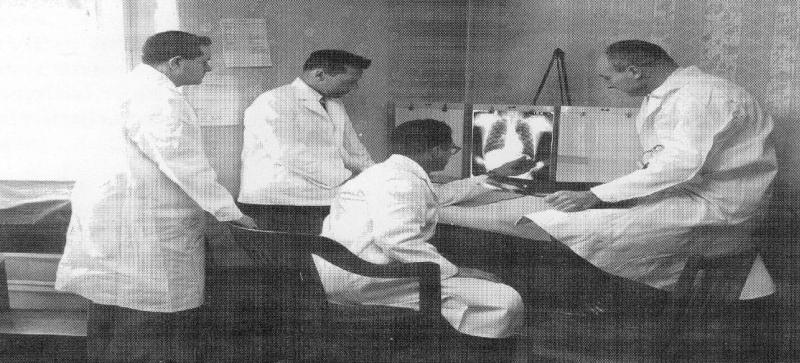 Four medical professionals in white coats examine X-ray images in a clinic, discussing findings to diagnose patients. The setting reflects early 20th-century medical practices.
