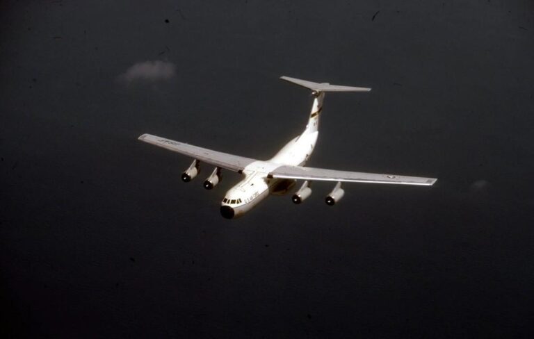 Military transport aircraft flying over a body of water, showcasing its wide wingspan and four engines against a dark sky backdrop.