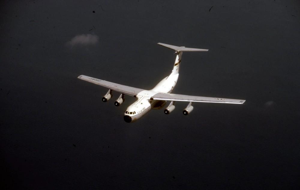 Military transport aircraft flying over a body of water, showcasing its wide wingspan and four engines against a dark sky backdrop.