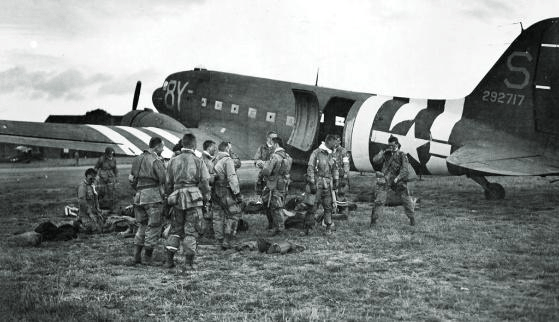Soldiers in World War II uniforms gather near a military aircraft, preparing for a mission. The backdrop includes a second plane with distinctive markings.