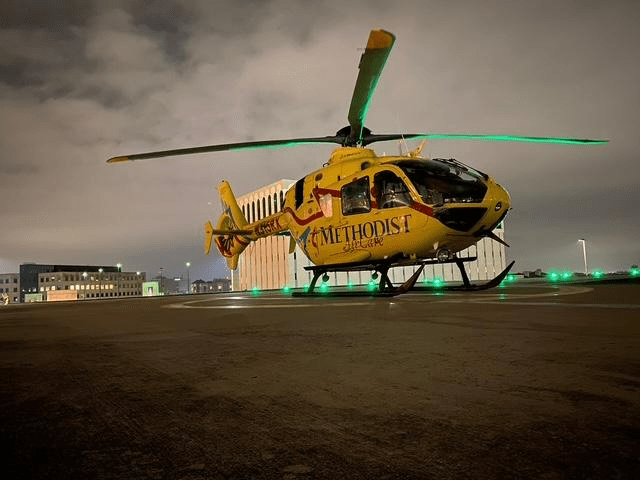 Yellow medical helicopter on a helipad at night, with city buildings in the background. It represents emergency medical services.
