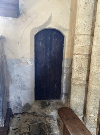 A weathered wooden door with an arched top set in a stone wall. The surrounding area features an old stone floor and wooden bench, suggesting historical significance.