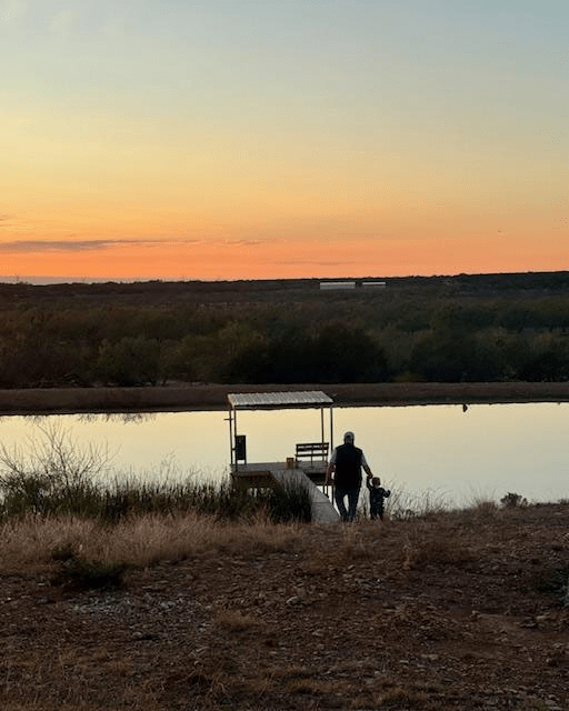 A person and a child walk toward a dock by a tranquil lake at sunset, surrounded by a natural landscape. The vibrant sky reflects serene outdoor leisure.
