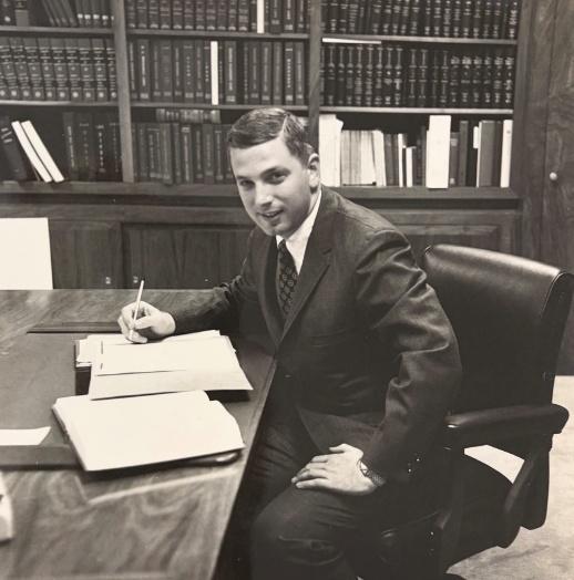 A young man in a suit sits at a desk, writing notes. Bookshelves filled with legal texts fill the background, suggesting a scholarly or professional setting.