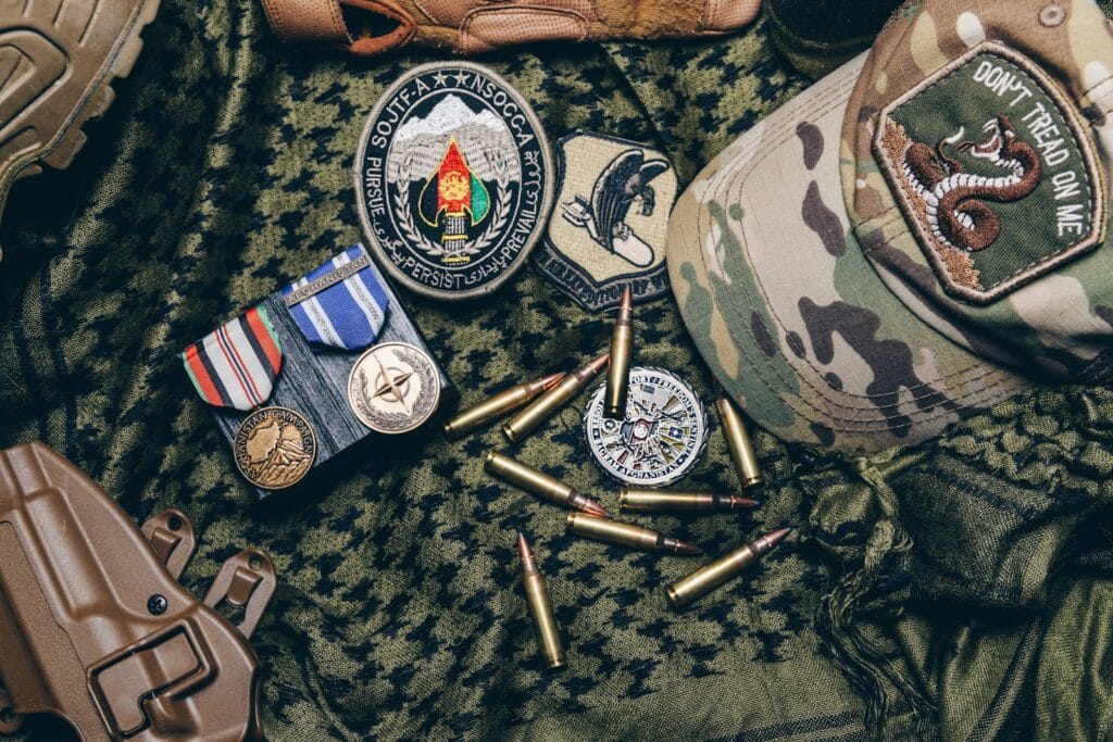 Military medals, patches, and a camouflaged hat are arranged on a tactical cloth, surrounded by ammunition, symbolizing service and readiness.